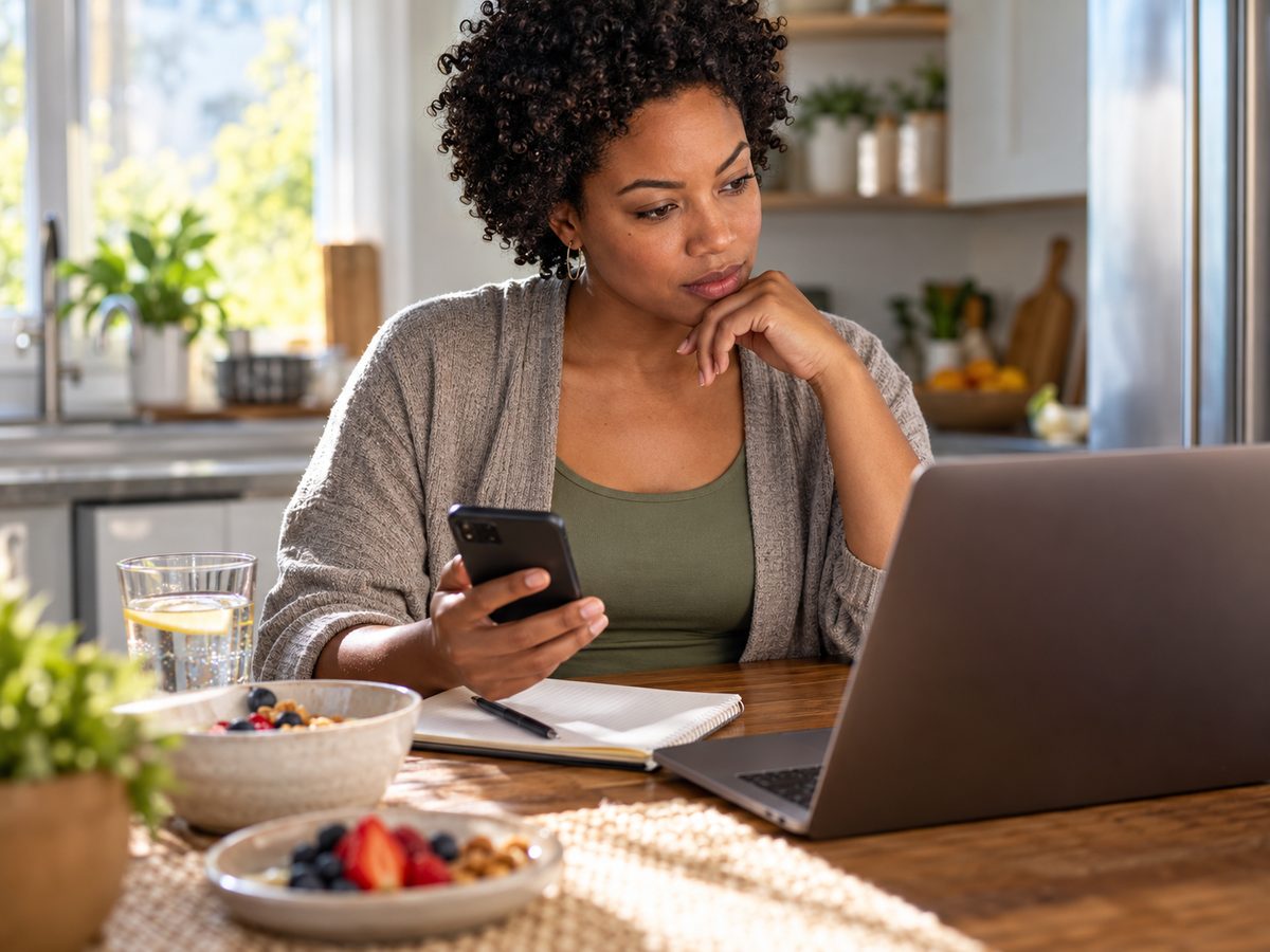 A woman at her kitchen table with her phone, laptop and notebook, planning her week.